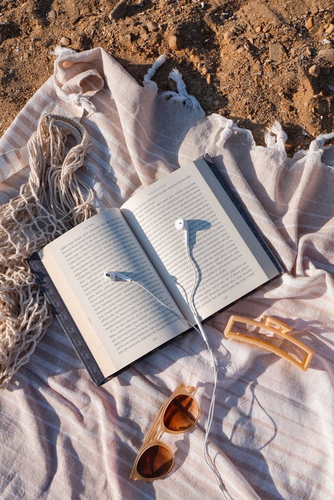 Top view of a beach setup with a book, sunglasses, and hair clip on a blanket.